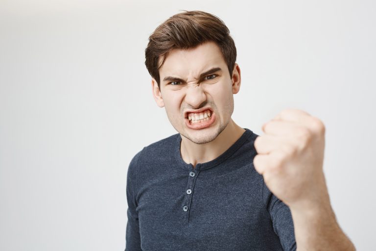 Fierce confident stylish european dark-haired male with trendy haircut holding fist in front of him as if ready for fight or challenge, clenching white teeth, having aggressive expression on face