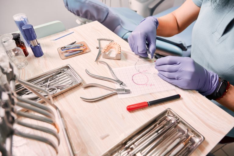 Close up view of female dentist in sterile gloves cutting braces wire while sitting at the table with orthodontic tools. Orthodontist preparing wire for braces attachment. Concept of dentistry.