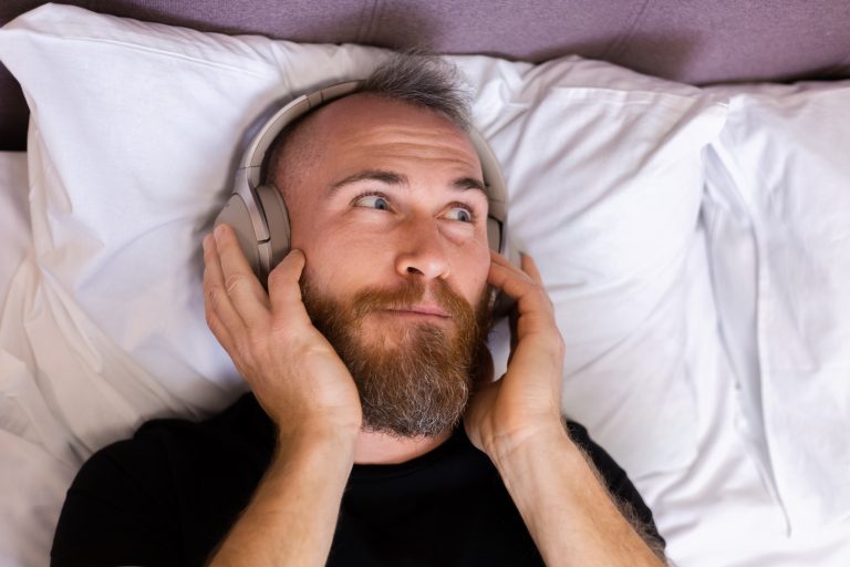 Happy caucasian man on bed wearing headphone listen enjoy his favorite music, resting alone, dancing.