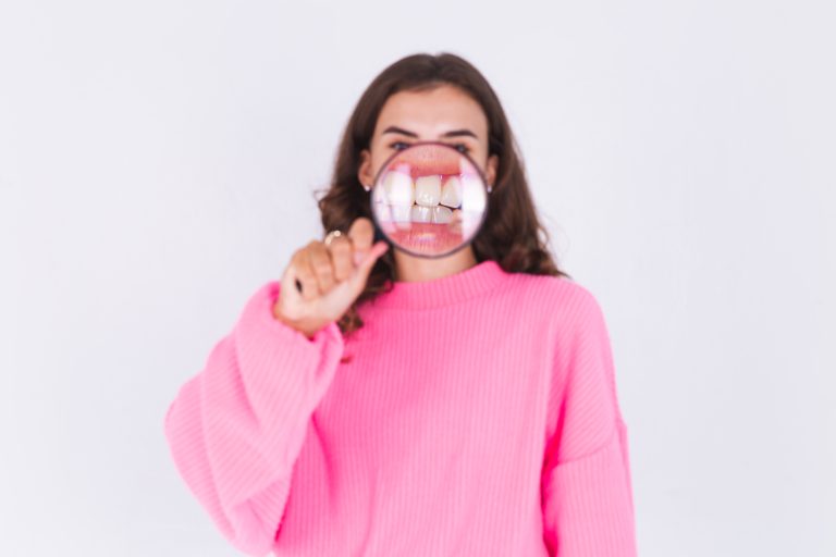 Young beautiful woman with freckles light makeup in sweater on white background with magnifier  shows white teeth perfect smile